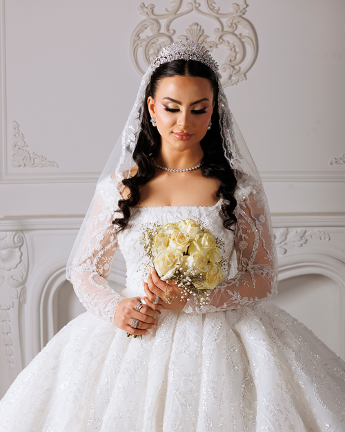 Bridal-Makeup-1 Bride in a white lace wedding gown holding a bouquet of flowers, standing outdoors with a soft background of trees and sunlight