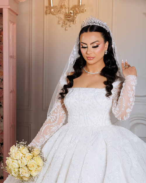 Bride in a white lace wedding gown holding a bouquet of flowers, standing outdoors with a soft background of trees and sunlight