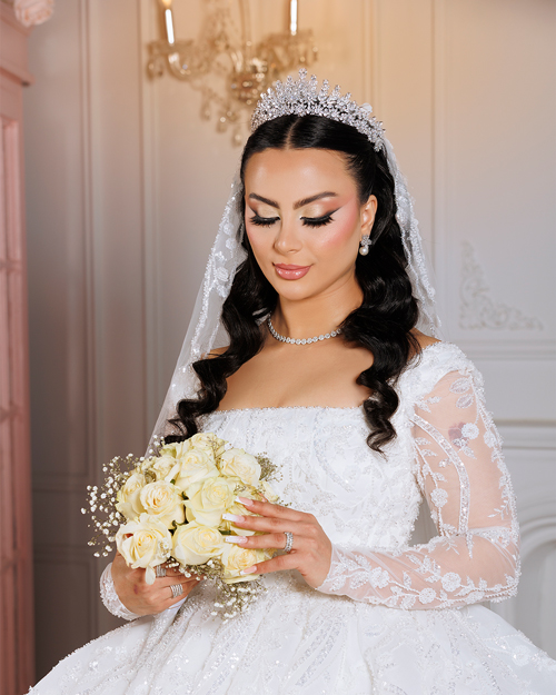 Bridal-Makeup-4 Bride in a white lace wedding gown holding a bouquet of flowers, standing outdoors with a soft background of trees and sunlight