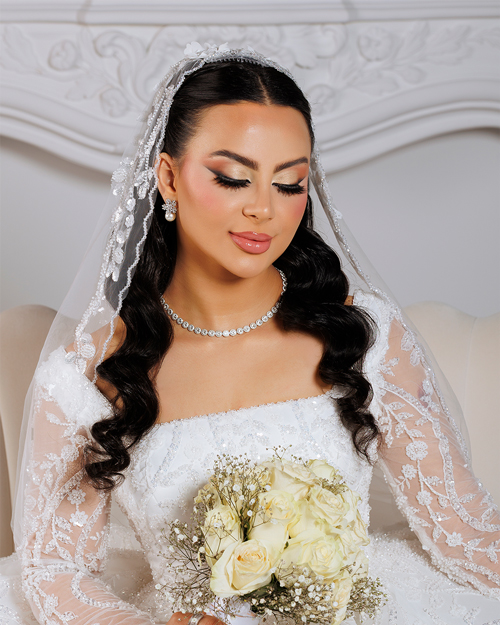 Bridal-Makeup-7 Bride in a white lace wedding gown holding a bouquet of flowers, standing outdoors with a soft background of trees and sunlight