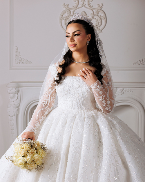 Bridal-Makeup Bride in a white lace wedding gown holding a bouquet of flowers, standing outdoors with a soft background of trees and sunlight