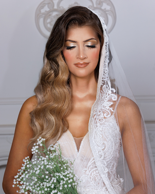 bridal_photo Bride in a white lace wedding gown holding a bouquet of flowers, standing outdoors with a soft background of trees and sunlight