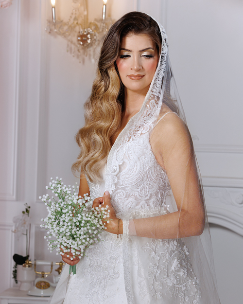 bridal_photo_2 Bride in a white lace wedding gown holding a bouquet of flowers, standing outdoors with a soft background of trees and sunlight