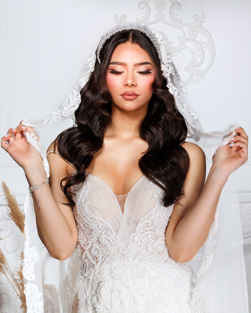bridal_photo_7 Bride in a white lace wedding gown holding a bouquet of flowers, standing outdoors with a soft background of trees and sunlight